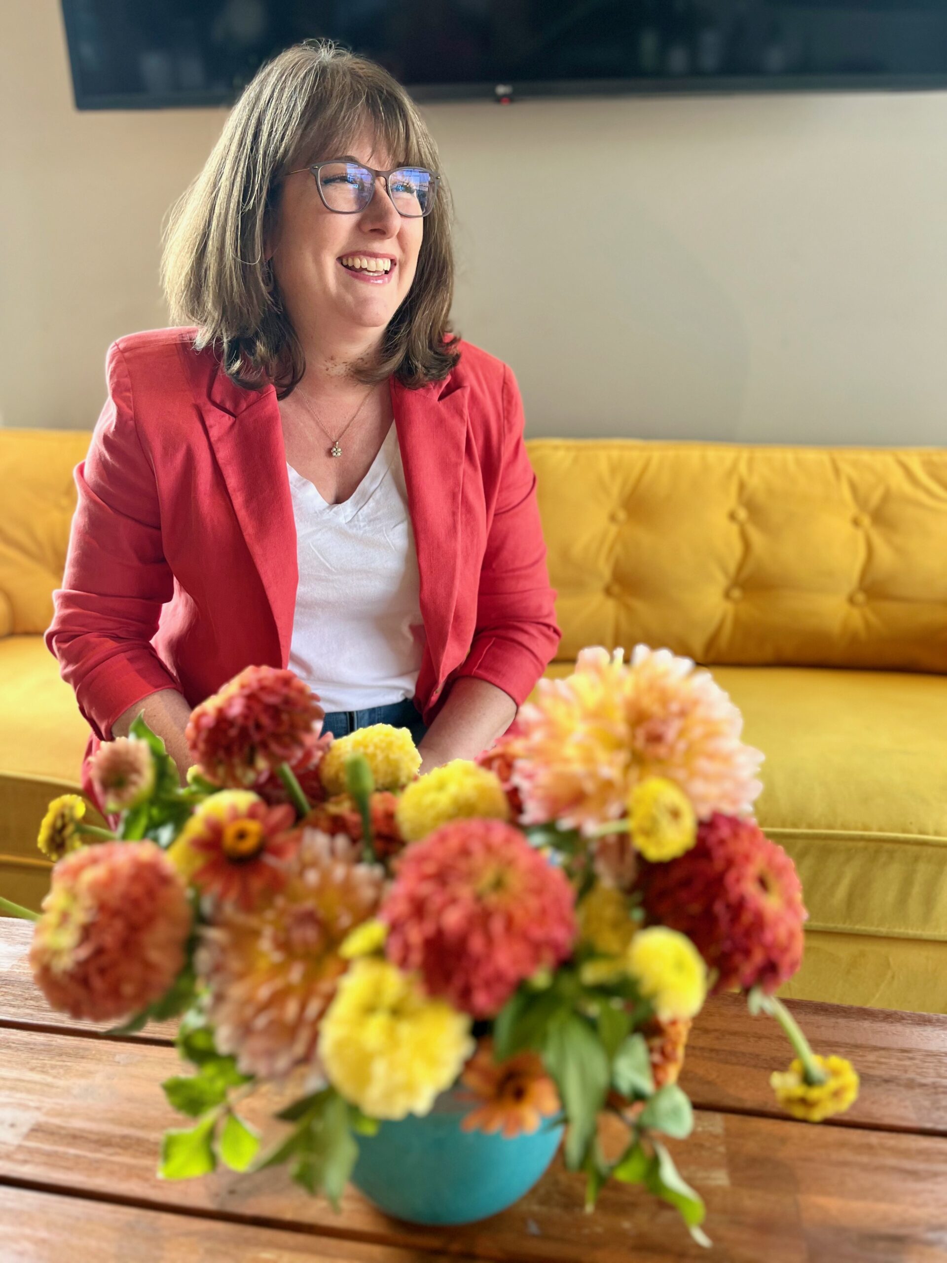 Becky Bivins seated on yellow couch wearing coral jacket next to colorful summer flowers